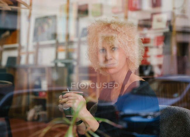 Através de vidro de artista feminina em avental sentado em poltrona no estúdio de arte e desfrutando de bebida no copo enquanto olha para a câmera — Fotografia de Stock