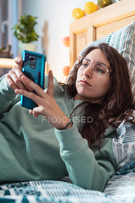 Молода жінка з бірюзовим светром і окулярами лежить на ліжку за допомогою мобільного телефону — стокове фото