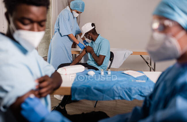 Vue latérale du médecin féminin en uniforme de protection et gants en latex désinfectant le bras avec du coton avec de l'alcool pour vacciner le patient afro-américain masculin en clinique pendant l'épidémie de coronavirus — Photo de stock