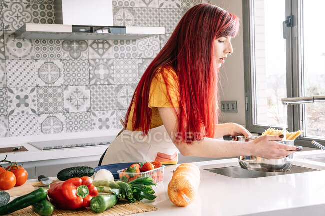 Молодая гомосексуальная женщина наливает воду из крана в кастрюлю с сырой пастой против разнообразных овощей в доме — стоковое фото
