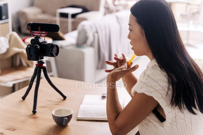 Side view of young ethnic female vlogger with notebook sitting at table with photo camera on tripod in kitchen — Stock Photo
