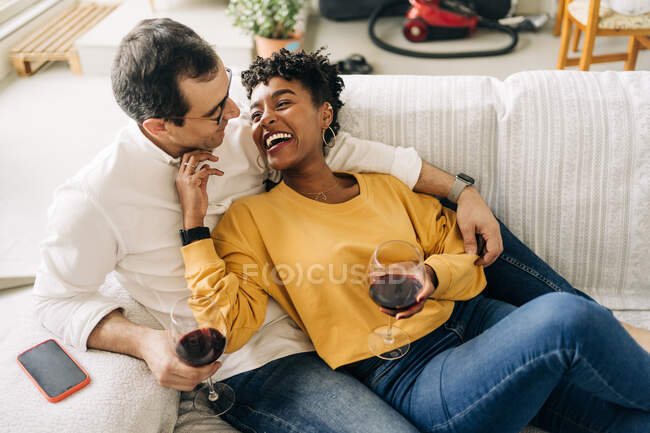 Conteúdo casal multirracial que arrefece no sofá em casa com vinho tinto em óculos enquanto desfruta de fim de semana em casa e olha um para o outro — Fotografia de Stock