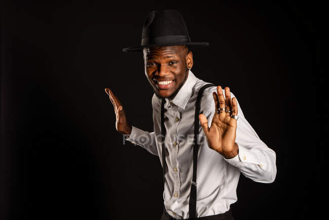 Young masculine ethnic male model in hat and trousers standing dancing while looking at camera on black background — Stock Photo