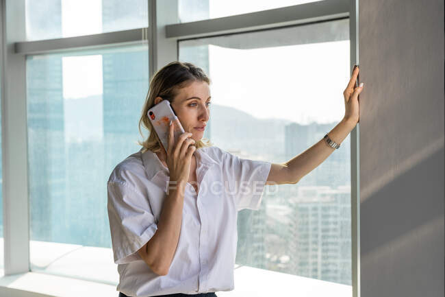 Молода бізнес-леді, стоячи в офісі з великими вікнами, має телефонний дзвінок на мобільний телефон — стокове фото