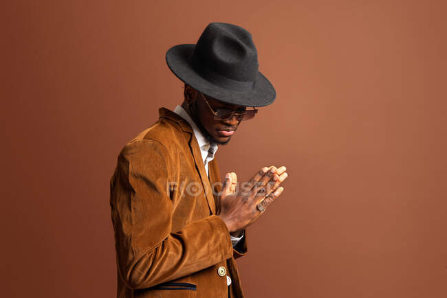 Vista lateral de un joven afroamericano con ropa y sombrero de moda mirando hacia otro lado sobre fondo marrón - foto de stock