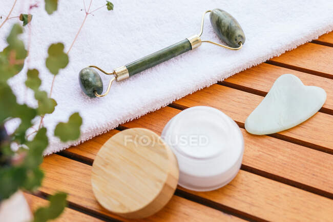 Alto ângulo de rolo de jade e raspador de guasha colocado na mesa de madeira com toalha e creme facial natural no salão de spa — Fotografia de Stock