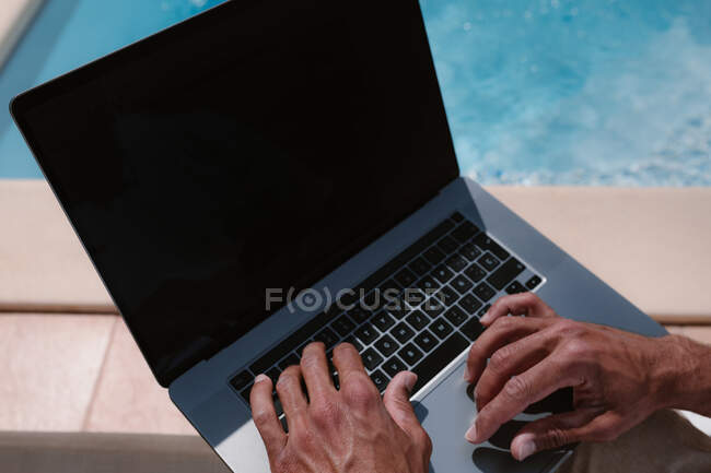 Cultivo irreconocible freelancer masculino acostado en la tumbona junto a la piscina y navegar por Internet en el ordenador portátil durante el teletrabajo en verano en el día soleado - foto de stock