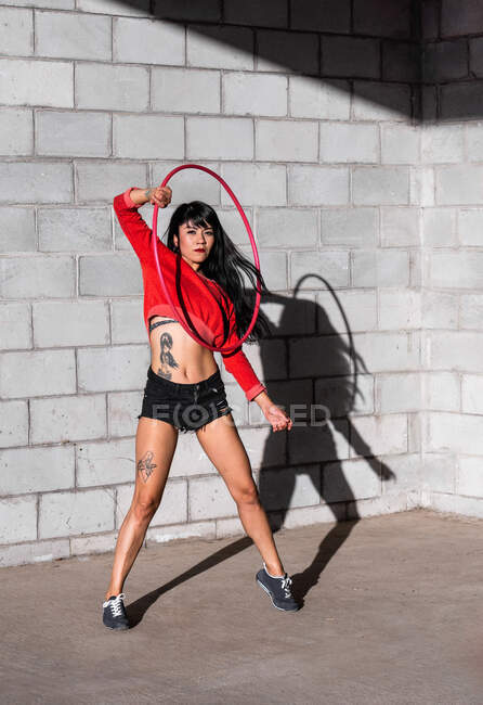 Young tattooed woman in activewear twirling hula hoop while dancing against brick walls with shadows and looking forward in sunlight — Stock Photo