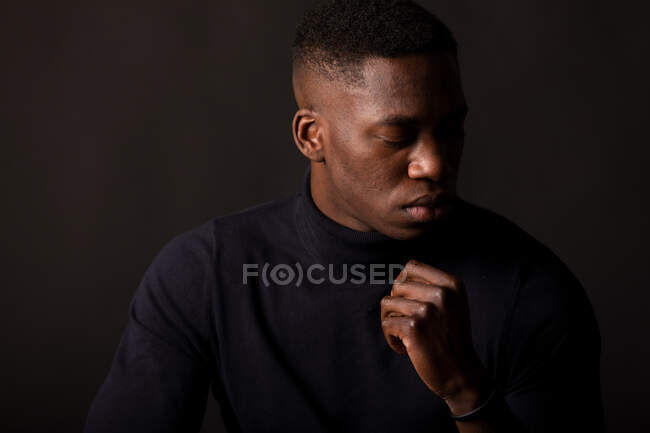 Confident young African American guy in black clothes with hand at chin closed eyes on black background in light studio — Stock Photo