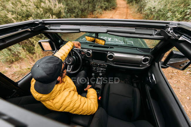 Vista interior desde arriba de un conductor con gorra y gafas de sol en un coche todoterreno mirando hacia otro lado - foto de stock