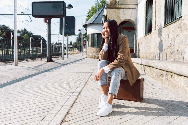 Delighted ethnic female traveler sitting on retro suitcase and leaning on hand while waiting for train at railway station and looking away — Stock Photo