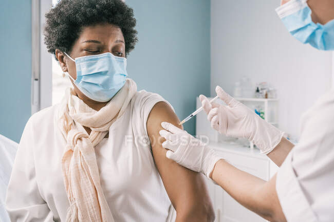 Especialista médica femenina irreconocible en uniforme protector, guantes de látex y mascarilla facial vacunando a una mujer madura afroamericana en la clínica durante el brote de coronavirus - foto de stock