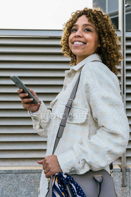 Mulher afro-americana otimista com penteado afro navegando no smartphone enquanto está contra a parede de metal na área urbana da cidade — Fotografia de Stock