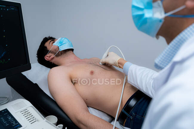 Побочный обзор обрезанного неузнаваемого мужчины-медика с помощью современного ультразвукового оборудования и обследование сердца пациента-мужчины в маске лежащего на медицинском столе во время диагностики в больнице — стоковое фото