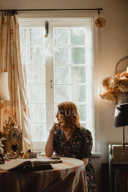 Jeune femme consciente regardant la table avec des livres contre la fenêtre dans la maison le jour ensoleillé — Photo de stock