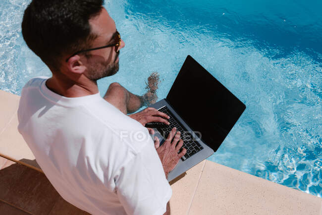Desde arriba, vista trasera del freelancer masculino en gafas de sol sentado junto a la piscina y navegando por netbook mientras trabaja de forma remota en el proyecto durante las vacaciones de verano - foto de stock
