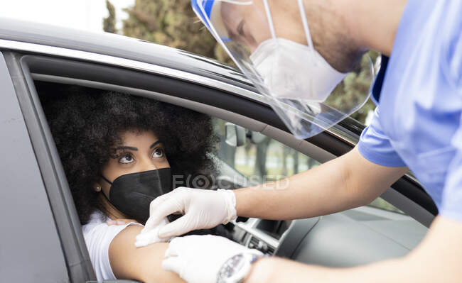 Male doctor in protective uniform, latex gloves and face shield disinfecting African American female patient arm with cotton and alcohol preparing to vaccinate inside the car on a drive through mobile clinic during coronavirus outbreak — Stock Photo