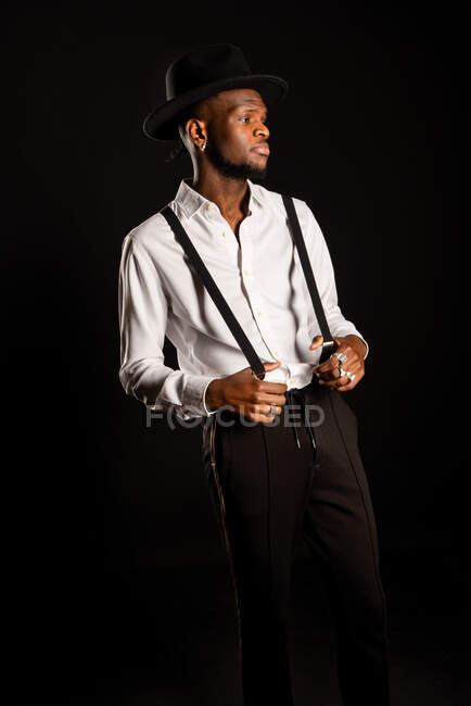 Joven modelo masculino masculino étnico en sombrero y pantalones de pie mientras mira hacia otro lado sobre fondo negro con humo - foto de stock