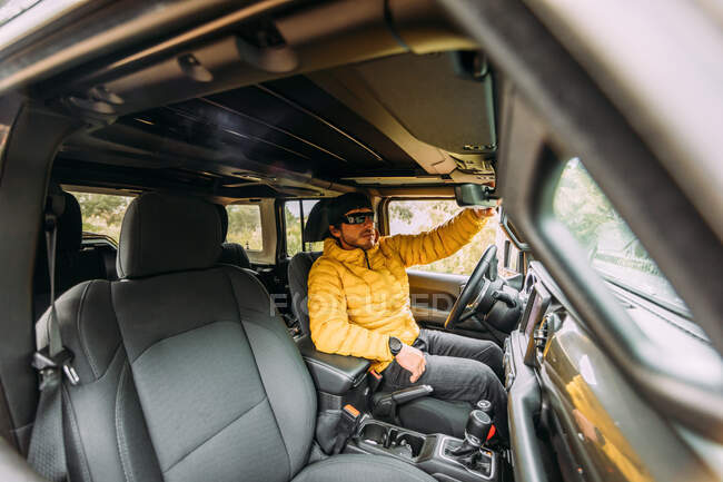 Inside view of a driver wearing a cap and sunglasses in an off-road car checking the internal rear view mirror — Stock Photo