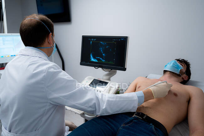 Чоловічий лікар з використанням сучасного ультразвукового обладнання та вивчення серця пацієнта у масці, що лежить на медичному столі під час діагностики в лікарні — стокове фото