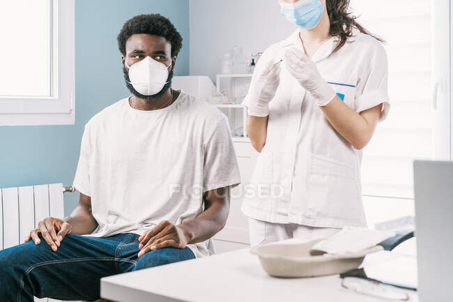 Обрезанная неузнаваемая женщина-врач в латексных перчатках и лицевом щите, наполняющая шприц из бутылки вакциной, готовящейся к вакцинации неузнаваемого пациента-афроамериканца в клинике во время вспышки коронавируса — стоковое фото