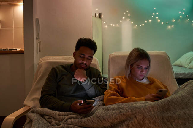 Спокойная многонациональная пара сидит под одеялом в уютной гостиной и просматривает мобильные телефоны во время отдыха дома в вечернее время — стоковое фото
