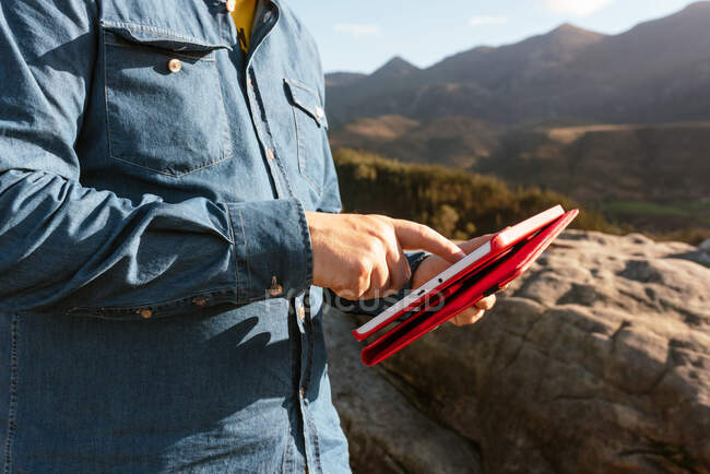 Vista lateral do explorador masculino tranquilo irreconhecível cortado com tablet sentado na rocha e desfrutando de uma vista espetacular das montanhas no dia ensolarado — Fotografia de Stock