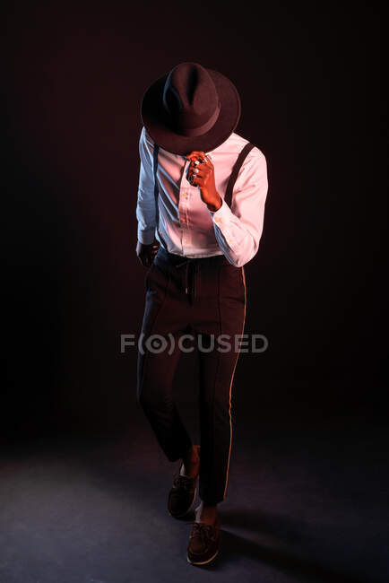 Joven modelo masculino masculino étnico en sombrero y pantalones de pie bailando mientras mira hacia otro lado sobre fondo negro - foto de stock