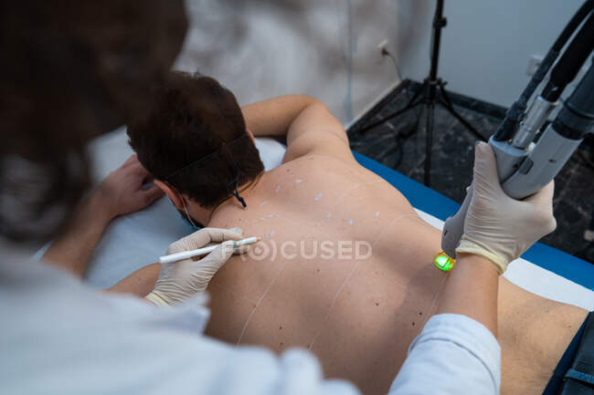 Maître méconnaissable utilisant le laser moderne sur le dos du client masculin pour l'épilation dans la clinique de beauté — Photo de stock
