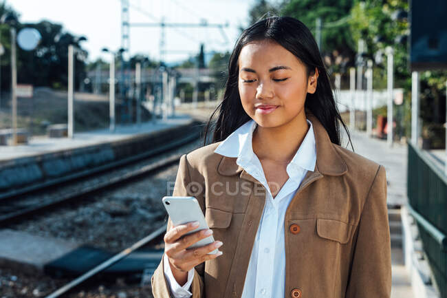 Cheerful Asian female traveler standing on platform at railway station and messaging on mobile phone — Stock Photo