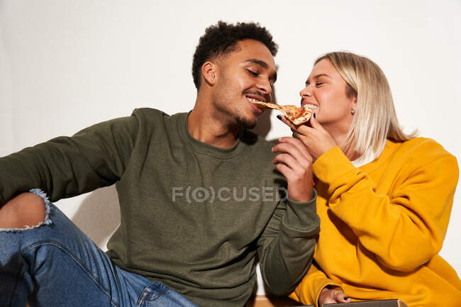 Позитивная многорасовая пара, поедающая пиццу вместе, развлекаясь и глядя друг на друга — стоковое фото