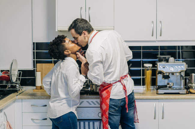 Вид збоку багатоетнічної пари в любові цілується, стоячи на кухні і готуючи — стокове фото