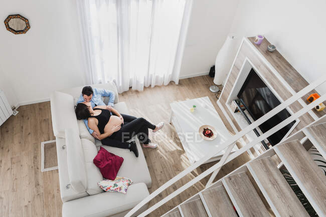 Desde arriba de la mujer embarazada sentada en el sofá con el hombre y besándose mientras disfrutan de fin de semana juntos en casa - foto de stock