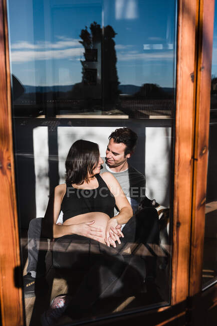 Através da janela da mulher grávida e do homem contente olhando ternamente um para o outro no dia ensolarado em casa — Fotografia de Stock