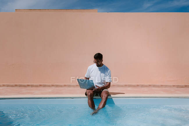 Freelancer masculino en gafas de sol sentado junto a la piscina y navegando por netbook mientras trabaja de forma remota en el proyecto durante las vacaciones de verano - foto de stock