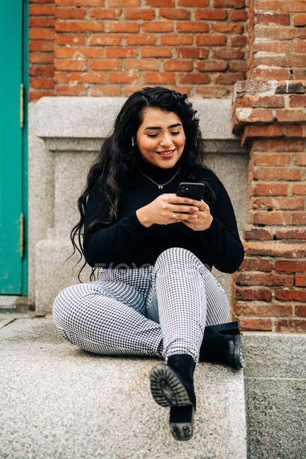 Восхитительный плюс размер женщины в модном наряде сидя на каменной границе в городе и обмена сообщениями на мобильный телефон — стоковое фото