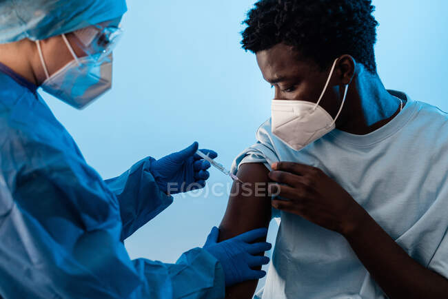 Female doctor in protective uniform and latex gloves vaccinating male African American patient in clinic during coronavirus outbreak — Stock Photo