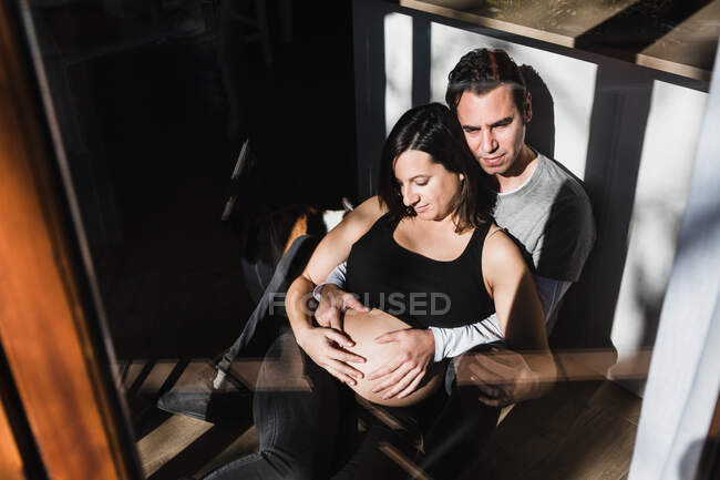 Через вікно зверху спокійний чоловік обіймає вагітну жінку ззаду і торкається живота, сидячи разом на підлозі в кімнаті, освітленій сонячним світлом — стокове фото