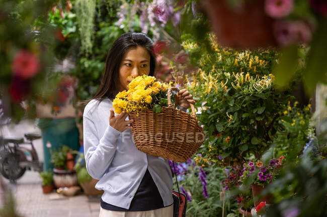 Belle fille asiatique achetant des fleurs dans la boutique de fleurs tout en portant un panier en osier avec des fleurs jaunes. — Photo de stock