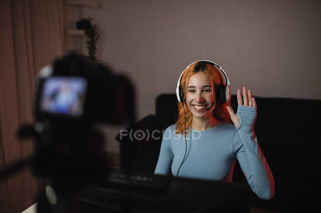 Sorrindo gamer feminino em fones de ouvido acenando a mão durante a gravação de vídeo na câmera profissional para blog de mídia social — Fotografia de Stock