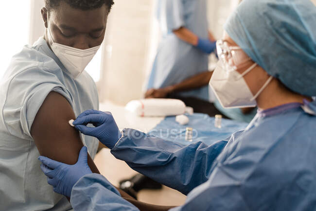 Visão lateral do médico feminino de uniforme protetor e luvas de látex desinfetando o braço com algodão com álcool para vacinar o paciente afro-americano masculino na clínica durante o surto de coronavírus — Fotografia de Stock