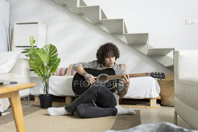 Young tattooed male guitarist sitting on floor and playing guitar at home — Stock Photo