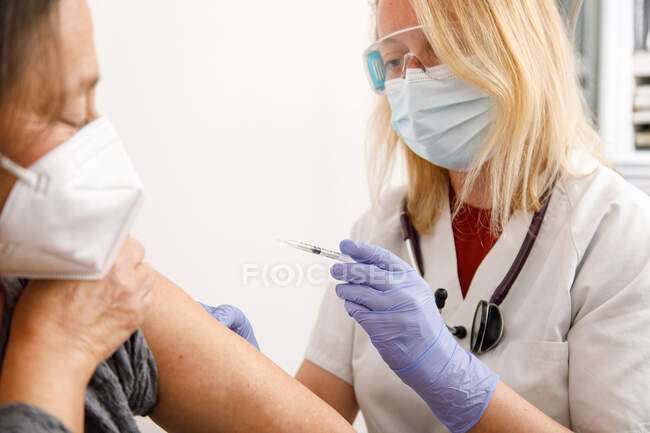 Spécialiste des cultures en uniforme de protection et gants de latex vaccinant une patiente âgée en clinique pendant l'éclosion de coronavirus — Photo de stock