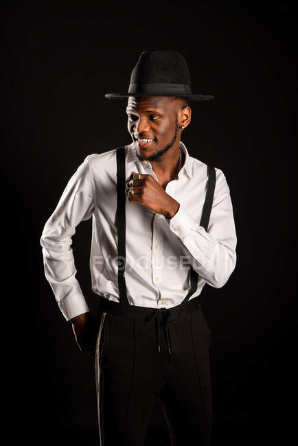 Joven modelo masculino masculino étnico en sombrero y pantalones de pie mientras mira hacia otro lado sobre fondo negro con humo - foto de stock