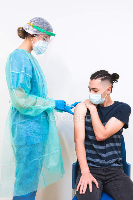 Especialista médica femenina en uniforme protector, guantes de látex y mascarilla facial vacunando a un paciente hispano en clínica durante brote de coronavirus - foto de stock