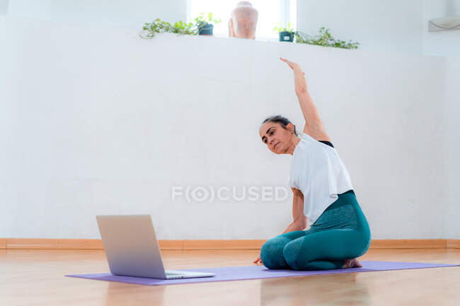 Mujer de mediana edad en ropa deportiva realizando inclinación lateral con brazo levantado mientras ve la lección de yoga en netbook en la habitación - foto de stock