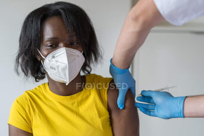 Spécialiste masculin méconnaissable en gants de latex protecteurs vaccinant une patiente afro-américaine en clinique pendant une épidémie de coronavirus — Photo de stock