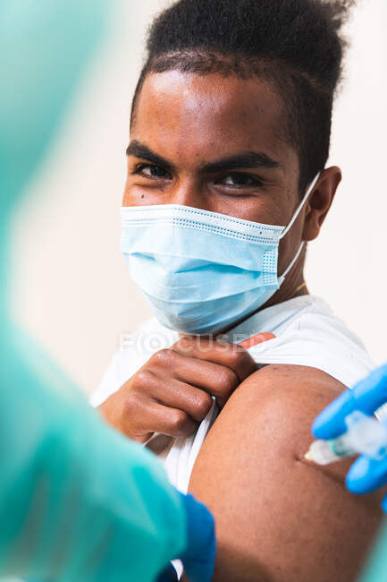 Especialista médica femenina irreconocible en uniforme protector, guantes de látex y mascarilla facial vacunando a un paciente afroamericano en la clínica durante el brote de coronavirus - foto de stock