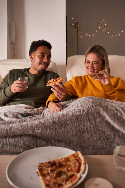 Многонациональная пара сидит в креслах и ест вкусную пиццу, наслаждаясь выходными вместе дома — стоковое фото
