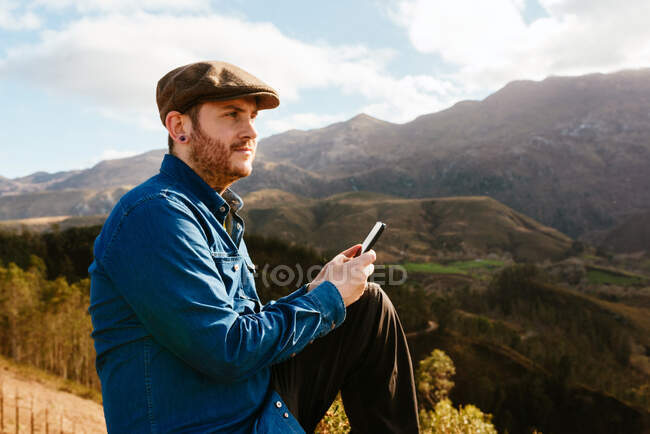 Vista lateral del caminante masculino sentado en la colina y el teléfono inteligente de navegación mientras admira el paisaje montañoso - foto de stock
